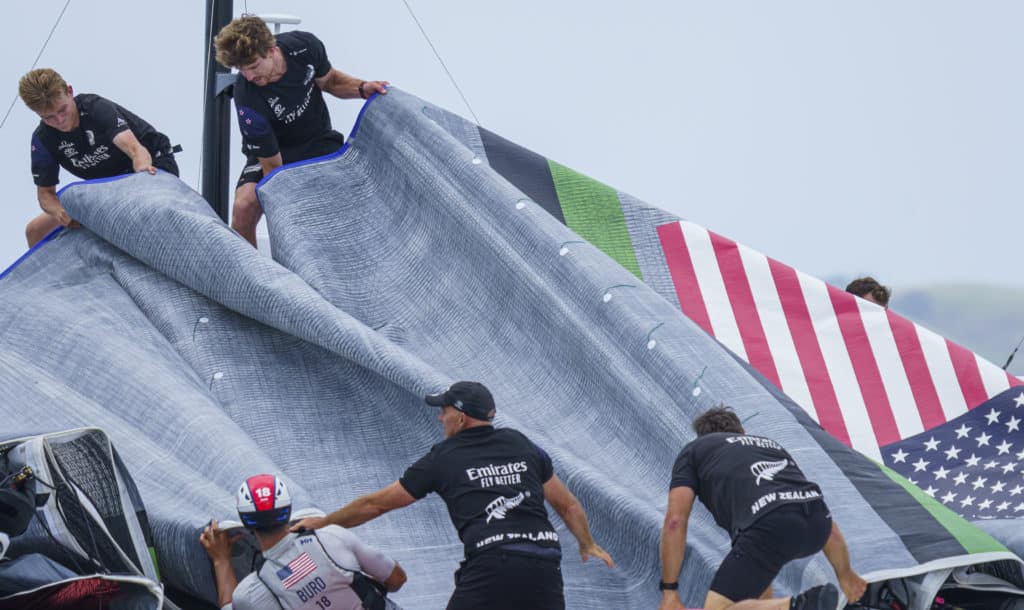 America's Cup sailors handling a mainsail with an American Flag logo.