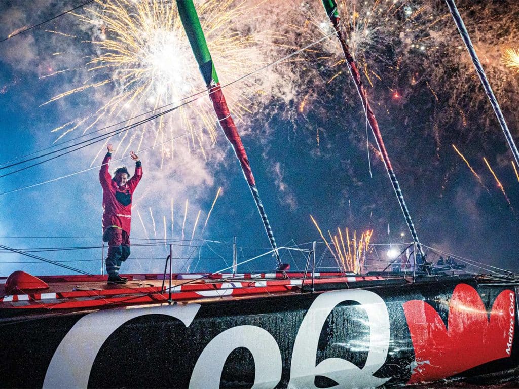 A man stands atop a sailboat with fireworks in the sky.